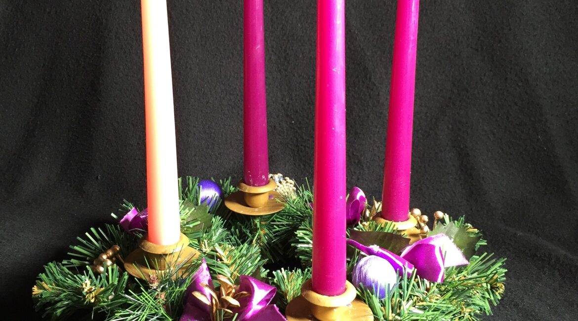 The four candles of the Advent wreath burn brightly. The wreath is shown against a black background.