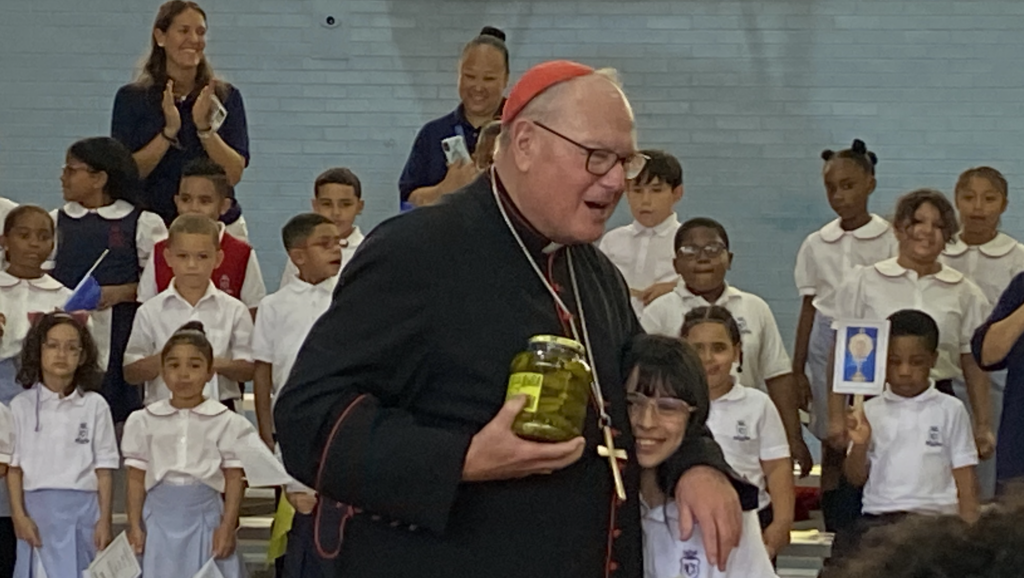 Cardinal Timothy Dolan embraces Holy Cross School fifth-grade student Isabella Colon after receiving three gifts, including a jar of pickles. The pickles are symbolic of Holy Cross School's original site in 1923, which was in a former pickle factory.