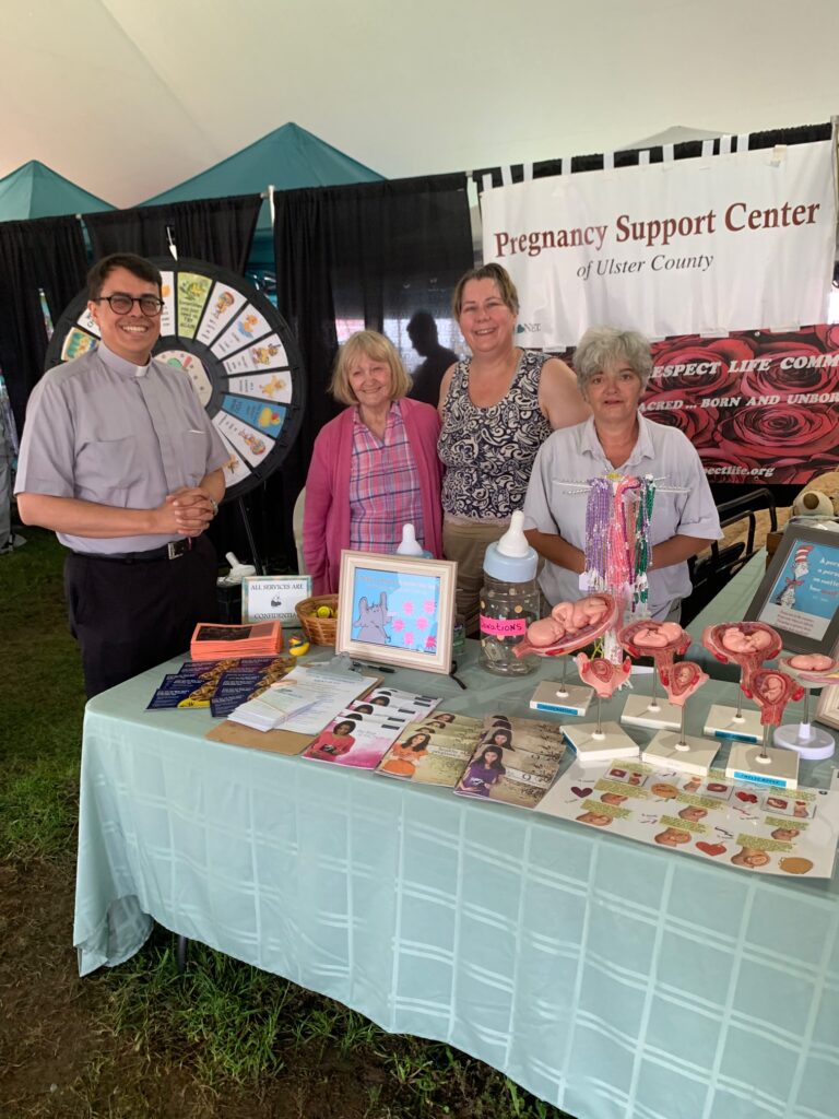 Father Arthur Rojas, Jane Randazzo (second left), and Catherine Parrinello (far right) join Michelle Whittaker (second right) at the joint booth of the Bravo Center and UDRLC.