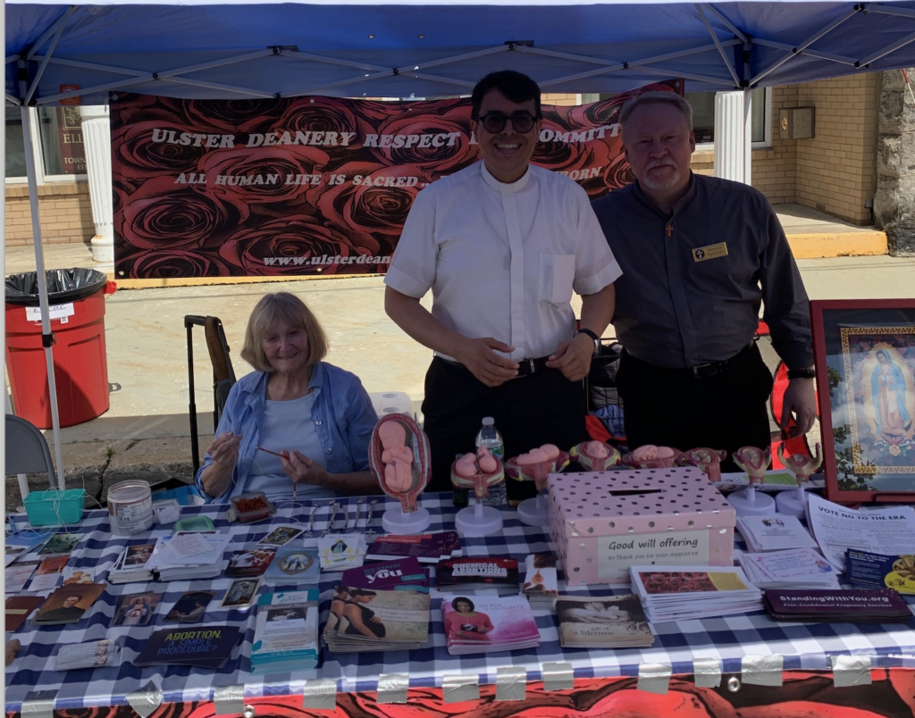 At the Ulster Deanery Respect Life Committee (UDRLC) booth on Canal Street in Ellenville, Jane Randazzo (seated) joins the Rev. Arthur Rojas, moderator and spiritual director of the UDRLC (center) and Deacon John Carr (right) in engaging visitors to the Blueberry Festival on a sunny August day in Ellenville.