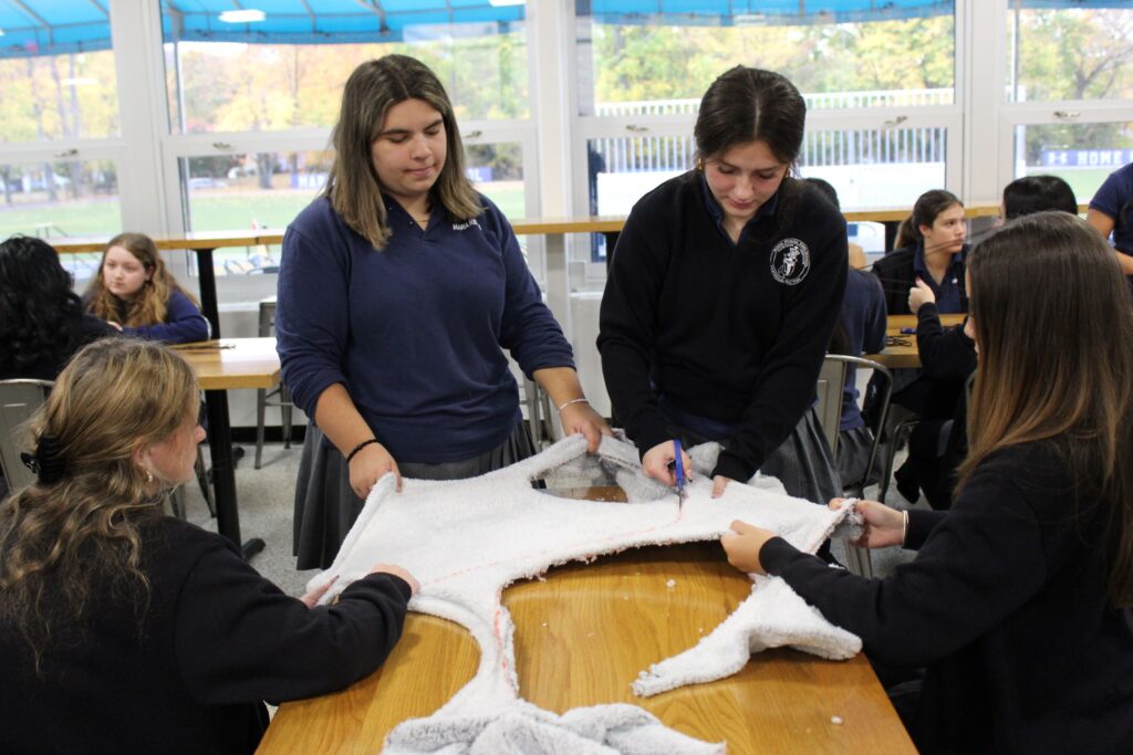 Maria Regina High School juniors (top, l-r) Olivia Maskara of the Bronx and Alexa Candelaria of Yonkers cut the fabric to craft the pouches that will be used for feminine hygiene products to support the young women of Tanzania.