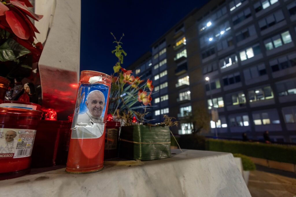 Votive candles, including some bearing a photo of Pope Francis, are seen on the base of a statue of St. John Paul II outside Rome's Gemelli hospital February 15, 2025.