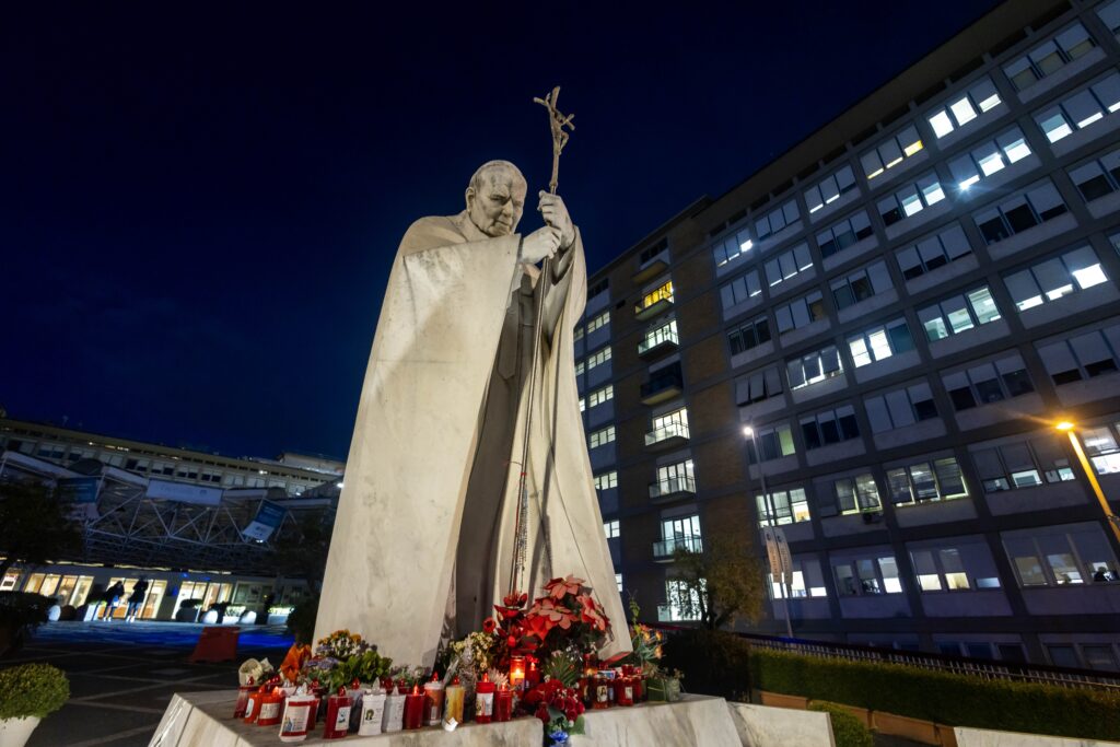 A statue of St. John Paul II, with flowers and candles at its base, is seen outside of Rome's Gemelli hospital in the early evening February 15, 2025, the day after Pope Francis was admitted for tests and treatment of a respiratory tract infection.