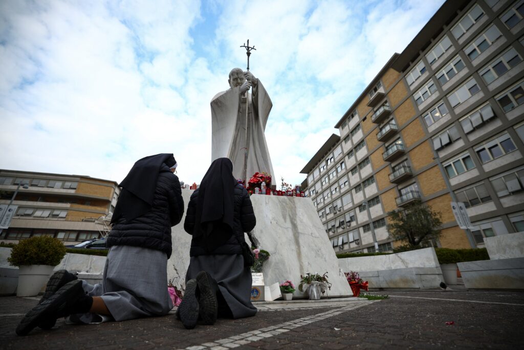 Monjas rezan junto a la estatua de San Juan Pablo II en el exterior del Hospital Gemelli de Roma el 20 de febrero de 2025, donde el Papa Francisco se encuentra ingresado para recibir tratamiento por una infección respiratoria.