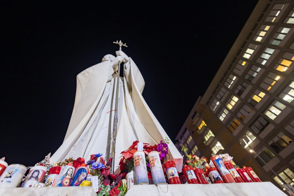 Votive candles and flowers are seen at the base of a statue of St. John Paul II outside Rome's Gemelli hospital February 21, 2025, where Pope Francis is being treated for double pneumonia.