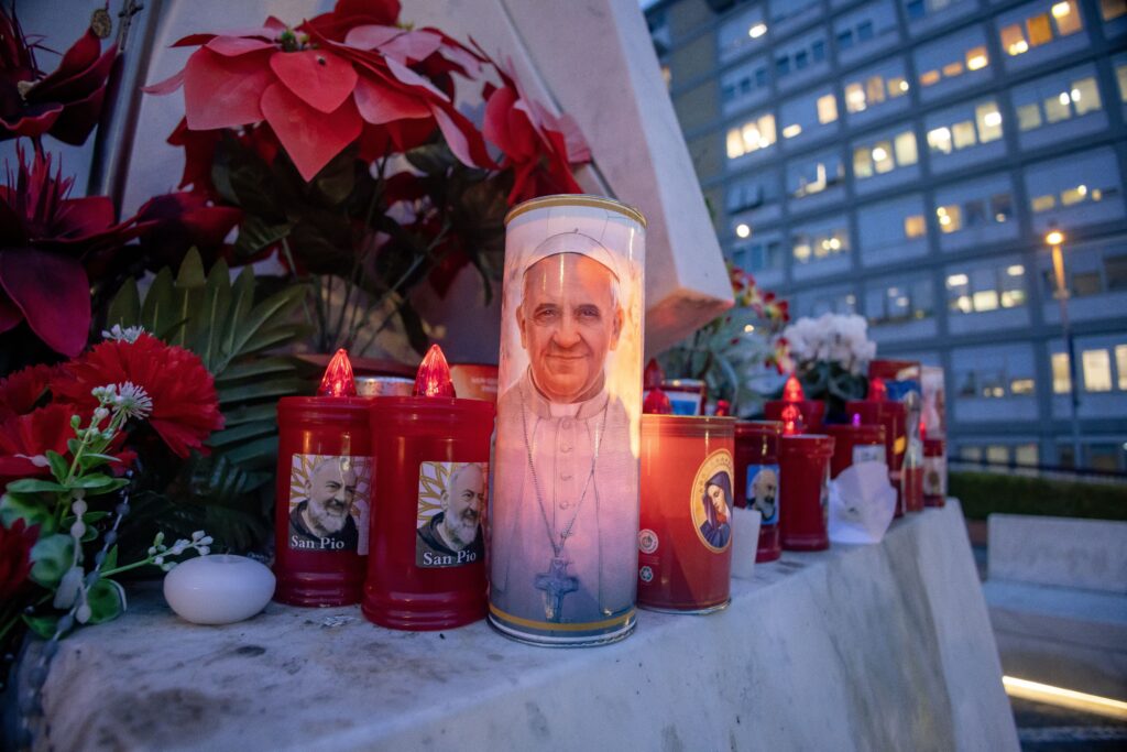 Velas votivas y flores se ven en la base de una estatua de San Juan Pablo II fuera del hospital Gemelli de Roma el 19 de febrero de 2025, donde el Papa Francisco está siendo tratado por una neumonía doble.