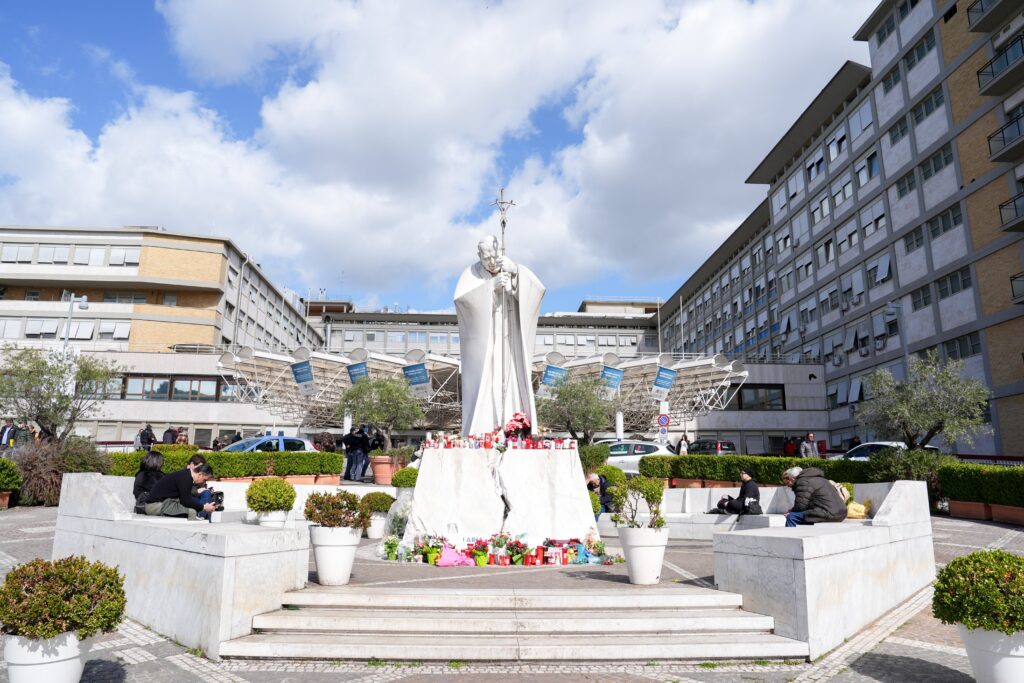 Velas votivas, flores y mensajes para el Papa Francisco se colocan en la base de una estatua de San Juan Pablo II afuera del hospital Gemelli de Roma el 27 de febrero de 2025, mientras el Papa Francisco recibe tratamiento por doble neumonía. (Foto CNS/Lola Gómez)