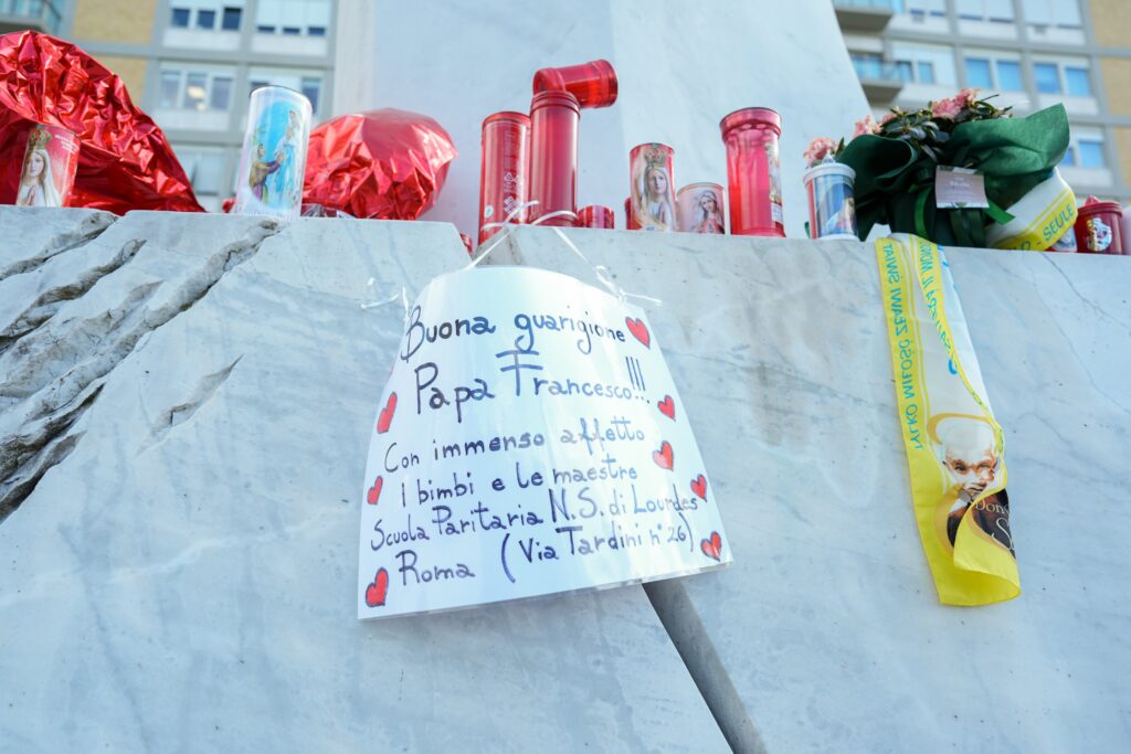 A handwritten sign with prayers for Pope Francis’ recovery is seen among candles and flowers at the base of a statue of St. John Paul II outside Rome’s Gemelli hospital February 27, 2025. (CNS photo/Lola Gomez)