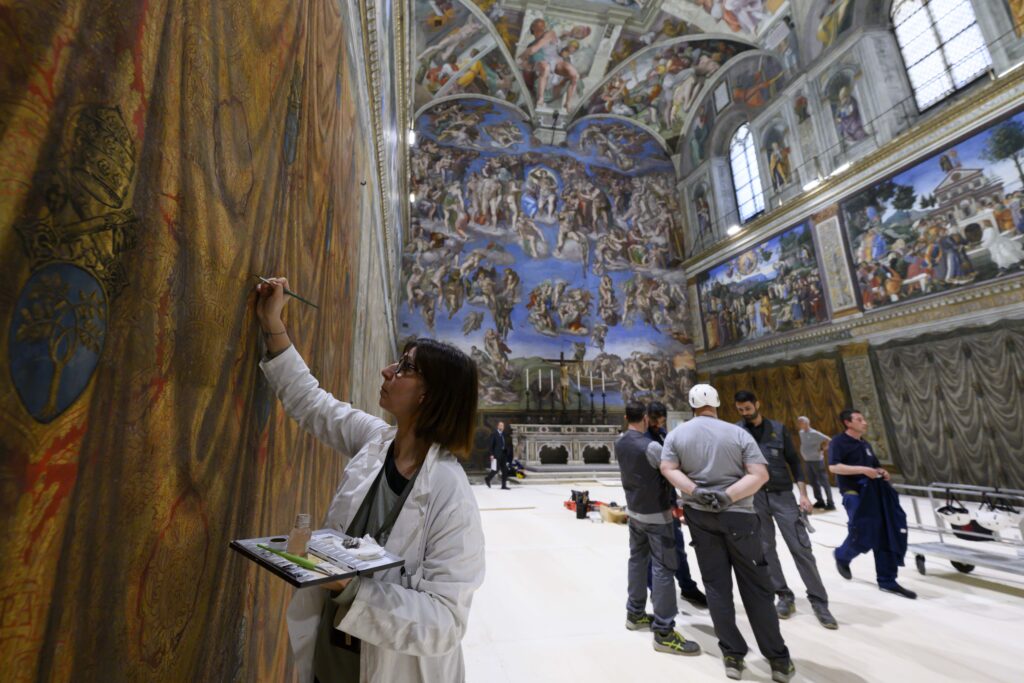 As a restorer with the Vatican Museums touches up a wall fresco in the Sistine Chapel at the Vatican May 2, 2025, workers prepare to build platforms over a protective covering on the marble floor May 2, 2025. The cardinals who enter the conclave to elect a new pope May 7 will sit at tables facing the center of the chapel with the tables in the back row raised slightly.