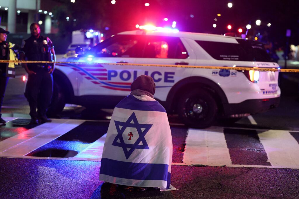 Un hombre, con una bandera israelí con una cruz en el centro, se arrodilla junto al personal de emergencias a primera hora del 22 de mayo de 2025, mientras trabajan en el lugar donde, según el Departamento de Seguridad Nacional de Estados Unidos, dos empleados de la embajada israelí fueron asesinados a tiros cerca del Capital Jewish Museum de Washington a última hora del 21 de mayo. El Comité Judío Americano estaba celebrando un evento nocturno en el museo.