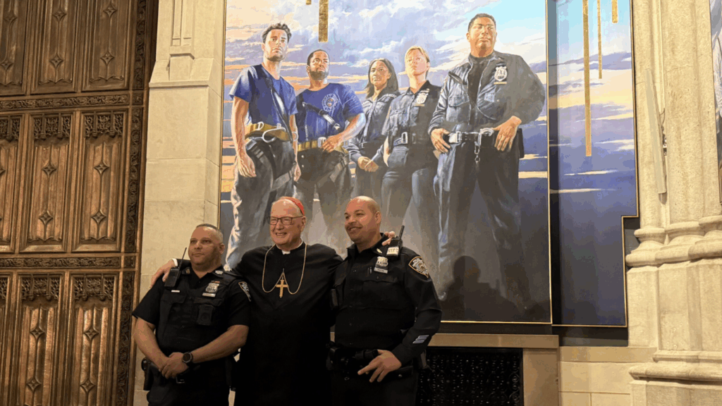 El cardenal Timothy Dolan (centro) posa con dos agentes del Departamento de Policía de Nueva York delante del panel dedicado a los rescatistas del nuevo mural de la catedral de San Patricio, “What's So Funny About Peace, Love, and Understanding” (¿Qué tiene de gracioso la paz, el amor y el entendimiento?), presentado en una rueda de prensa el jueves 18 de septiembre de 2025.