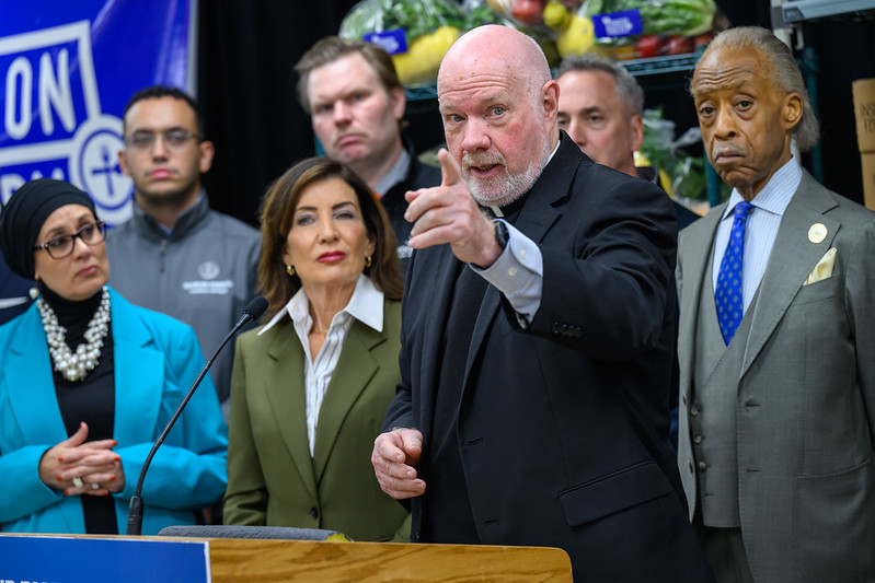 Monseñor Kevin Sullivan (centro) anuncia la respuesta de Caridades Católicas de Nueva York ante la próxima suspensión de los beneficios del SNAP, en una conferencia de prensa ofrecida por la gobernadora Kathy Hochul (centro izquierda) al declarar el estado de emergencia para abordar la situación.