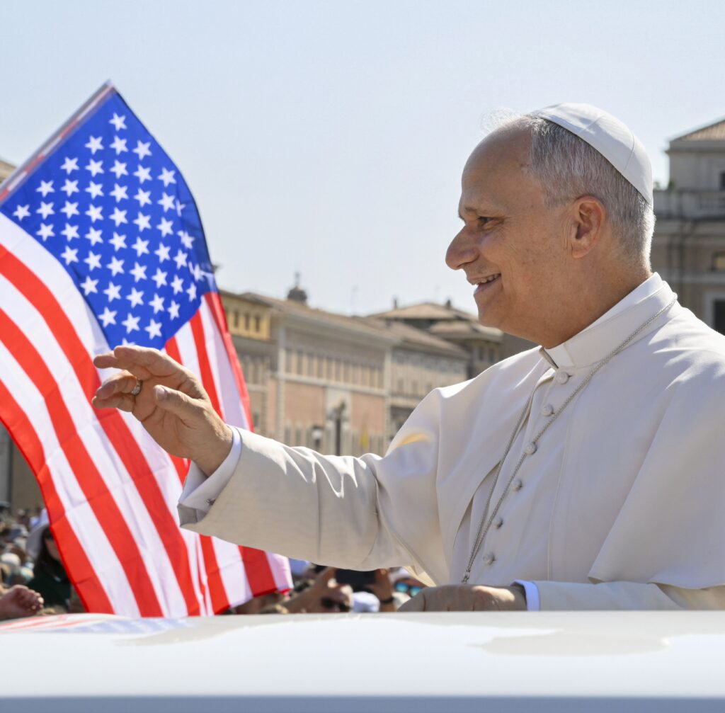 With a U.S. flag in the background, Pope Leo XIV waves to the crowd from the popemobile as he rides around St. Peter's Square at the Vatican before his weekly general audience on August 6, 2025.