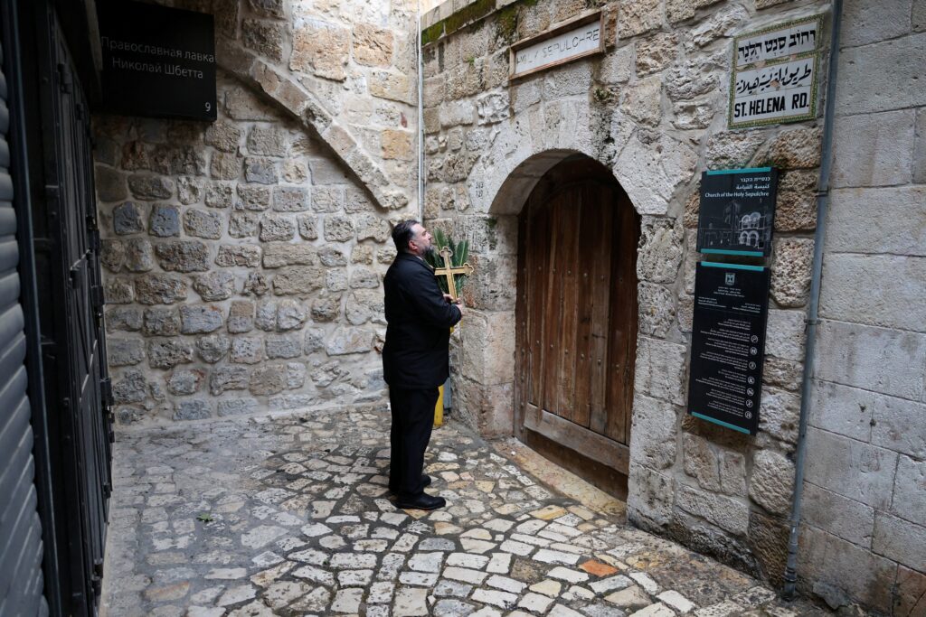 Issa Kassissieh, a Palestinian Christian who is popular for being the Santa Claus of the city, stands holding a cross and a palm frond at the doors of the Church of the Holy Sepulcher after finding them locked, following the cancellation of the traditional Palm Sunday procession from the Mount of Olives, amid restrictions on gathering in large groups and the U.S.-Israel war with Iran, in Jerusalem's Old City March 29, 2026.