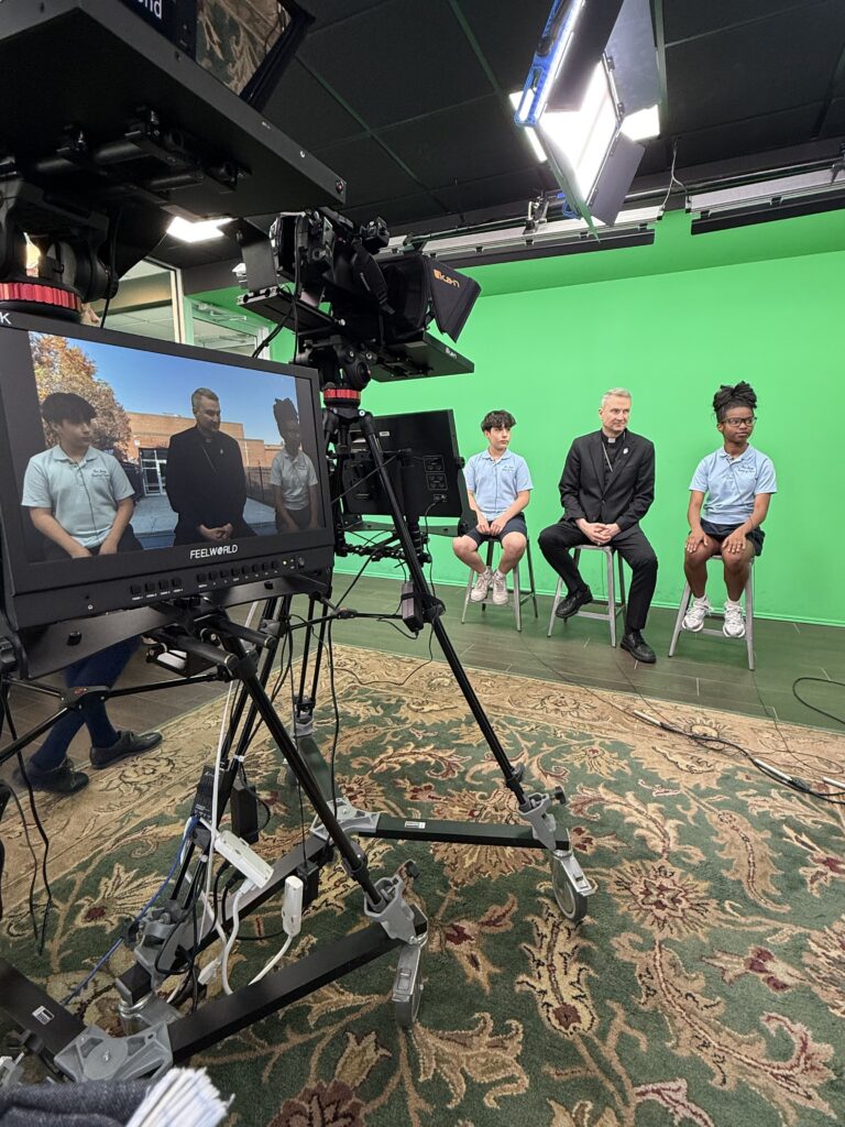 Angelo Barisciano (left) and London Ortiz (right of Archbishop Ronald Hicks), seventh grade students at our Lady Queen of Peace School, conduct an impromptu interview with Archbishop Ronald Hicks during his visit to Saint Joseph by-the-Sea High School, April 29, 2026.