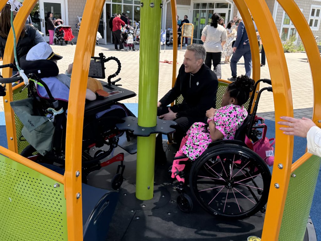 Archbishop Ronald Hicks (in black) rides a merry-go-round at Elizabeth Seton Children’s with the center’s international ambassador Stephanie and a resident, May 4, 2026.