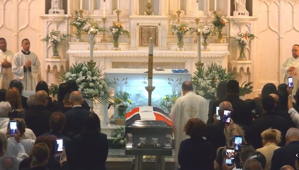 El reverendo Ambiorix Rodríguez, pastor de la Iglesia de St. Elizabeth, en Washington Heights, bendice el féretro donde yacen los restos del artista dominicano Alex Bueno, durante la Misa fúnebre del miércoles 24 de junio. Foto: captura de pantalla de la transmisión en vivo de Facebook de la Iglesia de St. Elizabeth