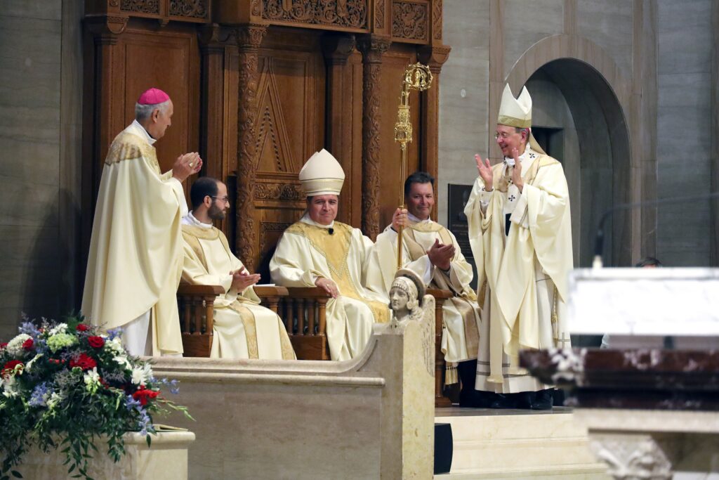 Bishop Evelio Menjivar-Ayala is seated in the cathedra, or bishop's chair, during his installation as the 10th bishop of Wheeling-Charleston, W.Va., July 2, 2026, at the Cathedral of St. Joseph in Wheeling. Archbishop Gabriele G. Caccia, apostolic nuncio to the U.S., left, Archbishop William E. Lori of Baltimore, right, and clergy look on, applauding.