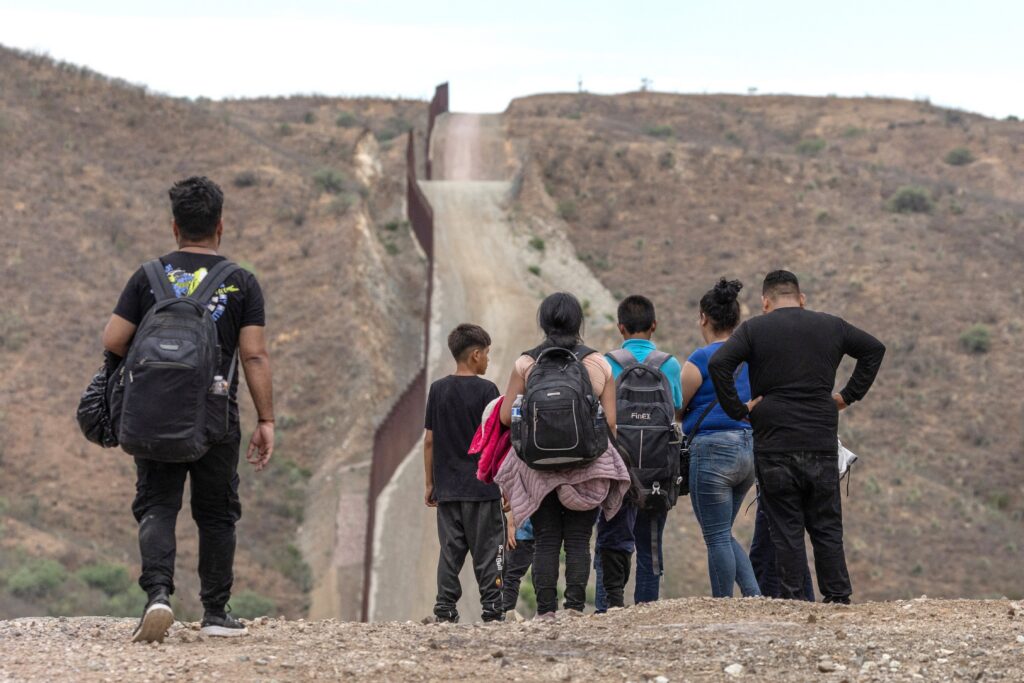 El muro fronterizo entre Estados Unidos y México se ve al fondo mientras migrantes de América del Sur y Centroamérica se preparan para entregarse a las autoridades de inmigración en Ruby, Arizona, el 24 de junio de 2024, tras cruzar a Estados Unidos desde México. Estrella del Paso, un ministerio católico de la Diócesis de El Paso, Texas, que atiende a decenas de miles de personas, ha agotado sus reservas tras siete meses sin recibir el pago del gobierno federal por su programa de representación de menores inmigrantes no acompañados.