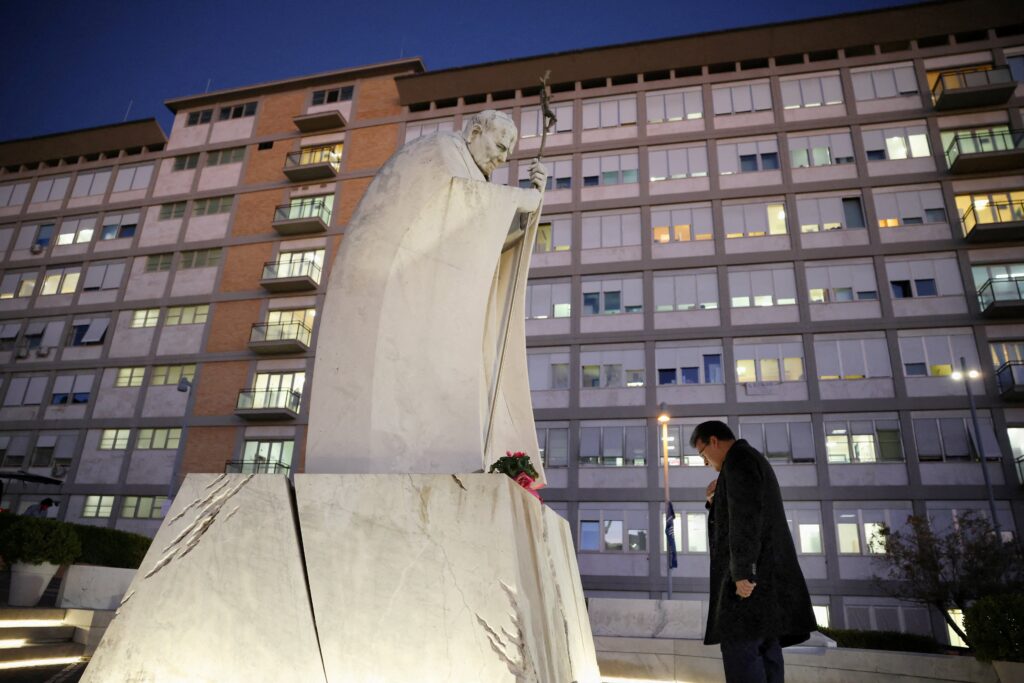 A man prays in front of a statue of the St. John Paul II outside Gemelli Hospital in Rome Oct. 13, 2026, where Pope Leo XIV had surgery that afternoon to remove a lung nodule. The Vatican confirmed that evening that Pope Leo has lung cancer.