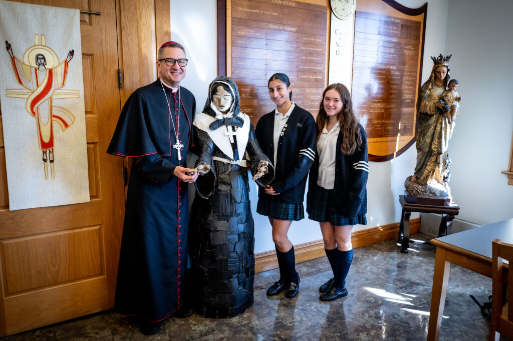 Archbishop Ronald A. Hicks stands with two Notre Dame Academy students and a statue of the school’s foundress, St. Marguerite Bourgeoys, on Oct. 22, 2026. The statue was handmade by the student out of cardboard and other upcycled materials.